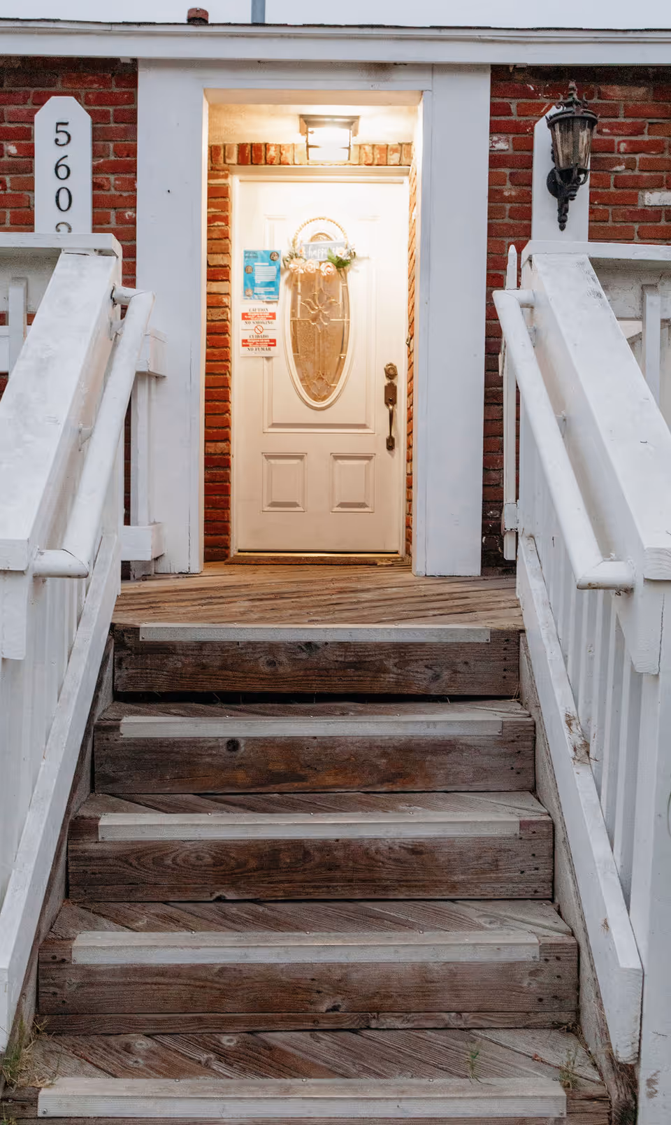 Front entrance of a building with wooden stairs leading up to a white door with an oval glass panel. The door is framed by red brick walls and white trim, with a light fixture above the door and a lantern-style light on the right side. The building number 5600 is displayed on a white vertical plaque to the left of the door.