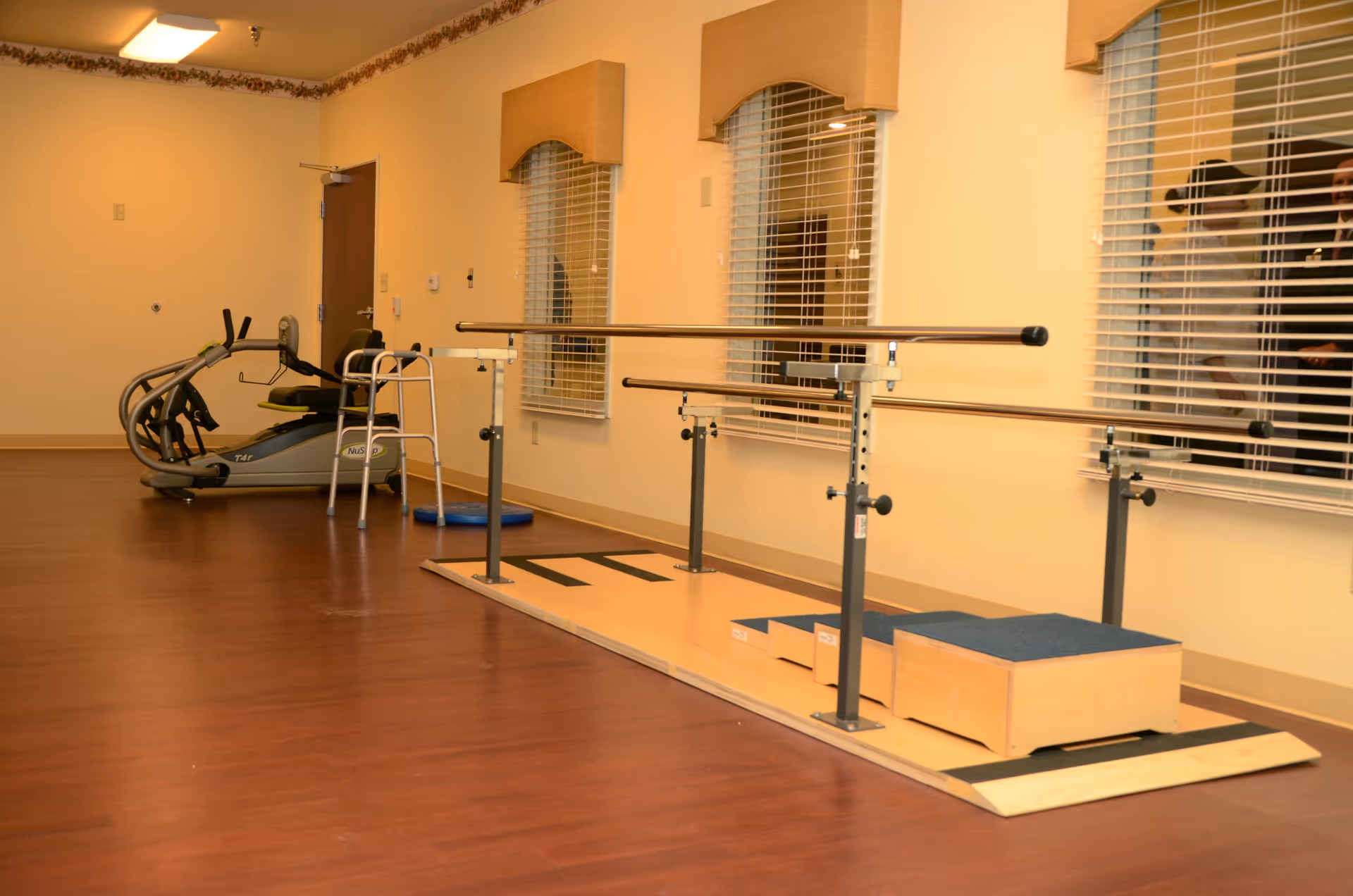 A rehabilitation or physical therapy room with parallel bars for walking exercises, a walker, an exercise bike, and wooden steps for mobility training. The room has wooden flooring, beige walls, three windows with blinds, and a door in the background.