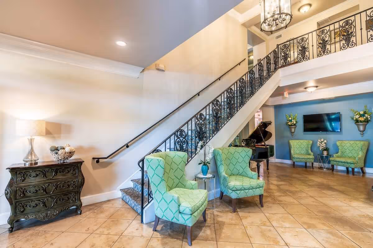 Well-lit lobby area with a decorative wrought-iron staircase, green upholstered chairs, a piano, and a side table with a lamp.