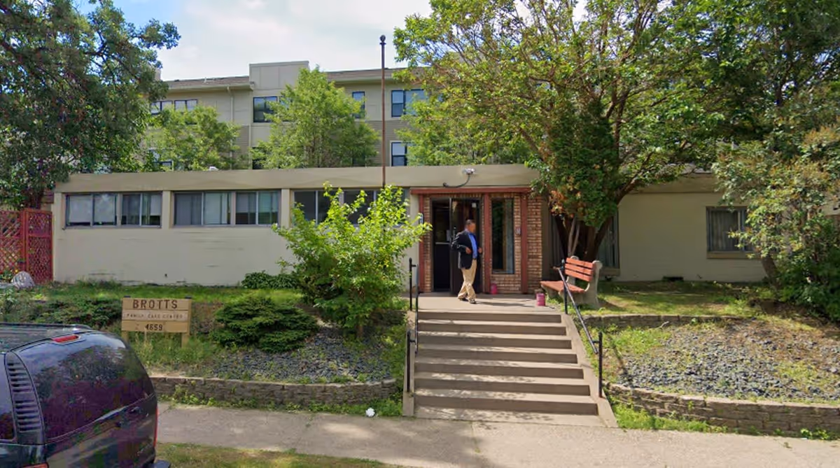 Exterior view of a single-story building with a staircase leading to the entrance. There is a person standing near the door, trees and bushes surrounding the building, and a parked vehicle partially visible on the left. A sign near the building reads 'BROTT'S Care Center 4659'.