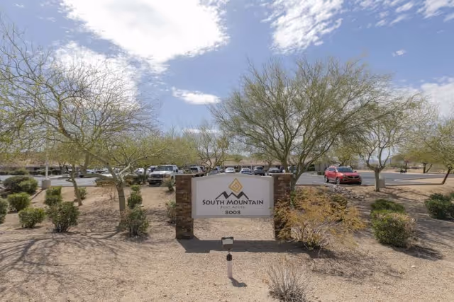 Outdoor view of the South Mountain Post Acute facility sign surrounded by desert landscaping with small trees and bushes, with a parking lot and several parked cars in the background under a partly cloudy sky.