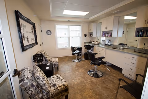 Interior view of a salon area in a senior living facility with two salon chairs in front of a counter with a sink and various hair care products. There are two patterned chairs along the left wall and a large window letting in natural light.