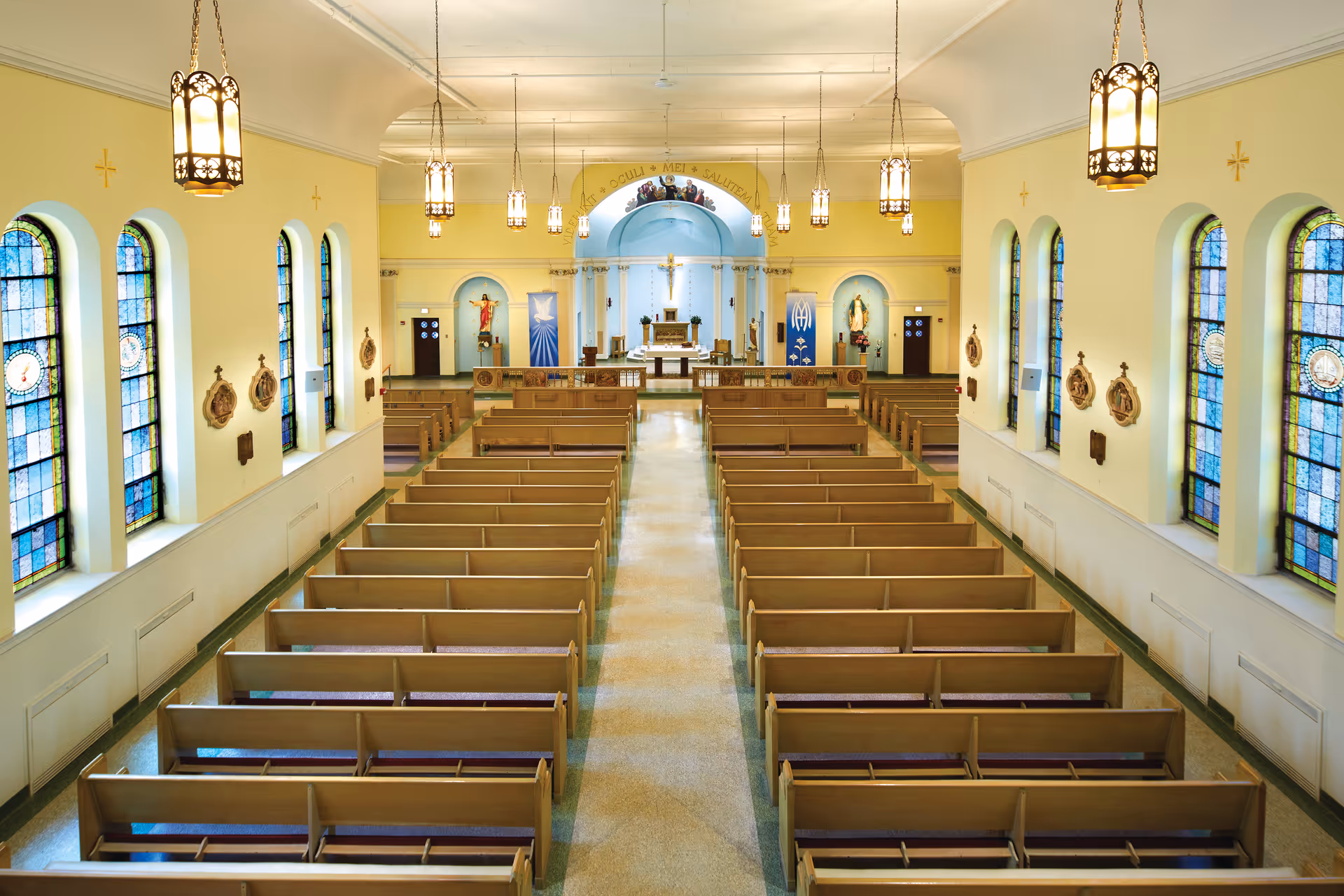 Interior view of a chapel with rows of wooden pews, stained glass windows on both sides, hanging lantern-style lights, and religious statues and banners near the altar at the front.