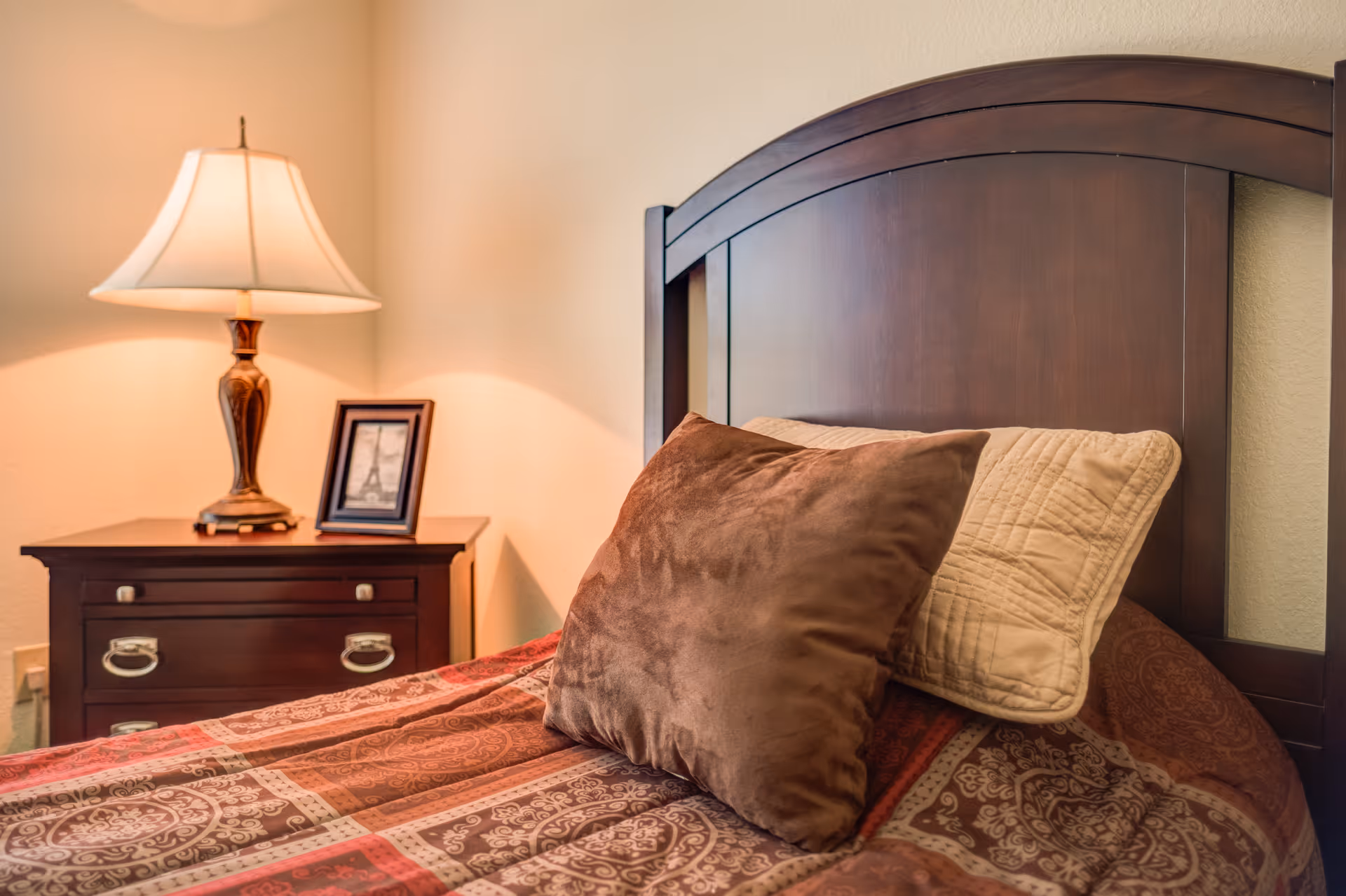 A cozy bedroom corner featuring a dark wooden headboard with two pillows, one brown and one beige, on a patterned bedspread. Next to the bed is a wooden nightstand with two drawers, a decorative table lamp, and a framed picture of the Eiffel Tower.