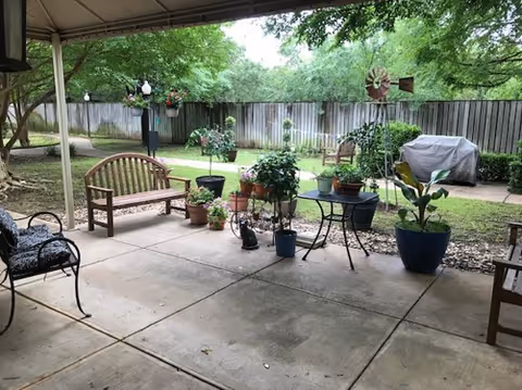 Covered patio with benches, potted plants, a small table, and a lawn with a wooden fence and grill in the background.