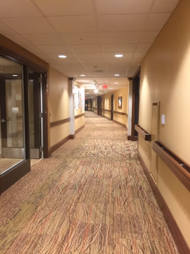 A long, carpeted hallway in a senior living facility with beige walls, wooden trim, handrails on both sides, recessed ceiling lights, and framed artwork on the walls. There is a glass door on the left side near the foreground.