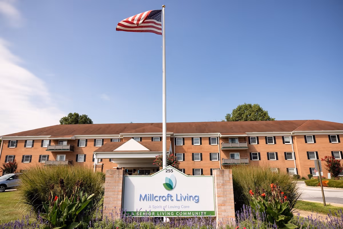 Exterior view of Millcroft Living senior living community building with a large American flag on a flagpole in front. The building is a three-story brick structure with balconies and windows. There is a sign in front with the facility name and slogan, surrounded by landscaping and flowers under a clear blue sky.