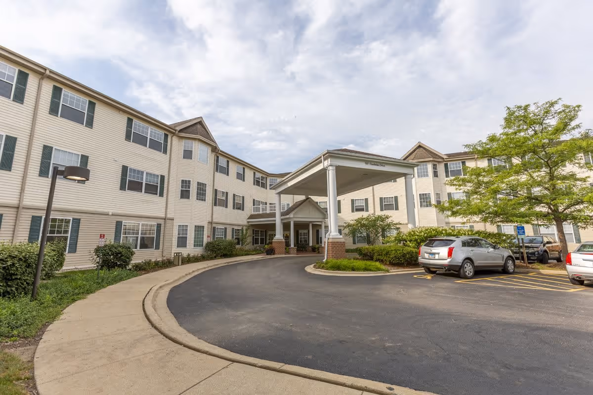 Exterior view of a three-story senior living facility building with beige siding and green shutters. There is a covered entrance with white columns and a circular driveway in front. Several cars are parked in the parking lot, and there are trees and shrubs around the building. The sky is partly cloudy.