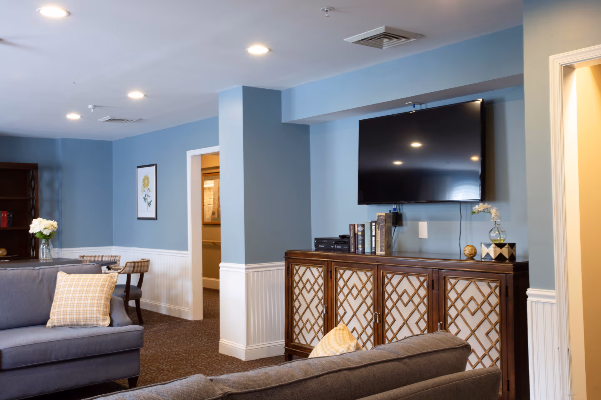A cozy living room with blue walls and white wainscoting. The room features two gray sofas with plaid pillows, a wooden cabinet with a lattice design, a flat-screen TV mounted on the wall above the cabinet, and a small vase with flowers on top. There is a framed floral picture on the wall and a small table with chairs in the background.