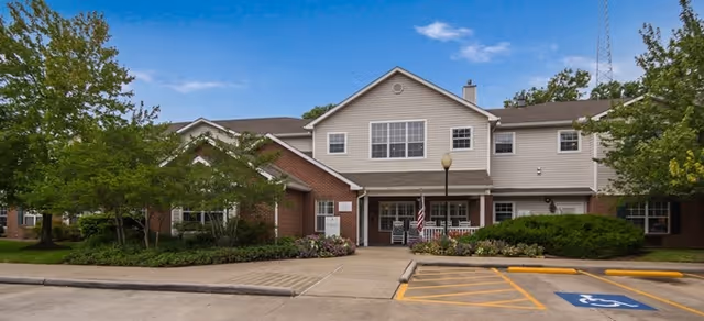 Exterior front view of a two-story senior living facility building with a combination of brick and beige siding. There is a paved driveway and parking area in front, including a handicapped parking space. The building is surrounded by green trees and shrubs under a clear blue sky.