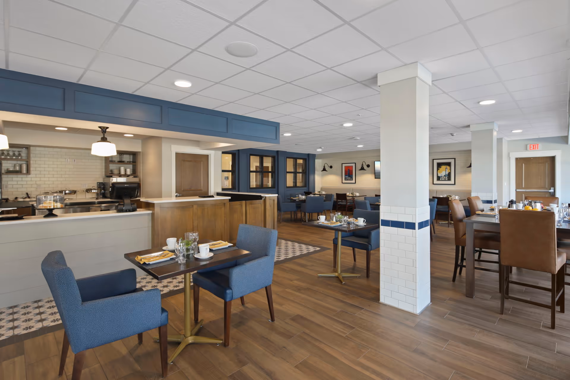 A spacious dining room in a senior living facility featuring wooden floors, multiple tables set with cups, plates, and napkins, blue upholstered chairs, and a counter area with a coffee machine and display case. The room has white tiled pillars with blue accents, framed artwork on the walls, and recessed ceiling lights.