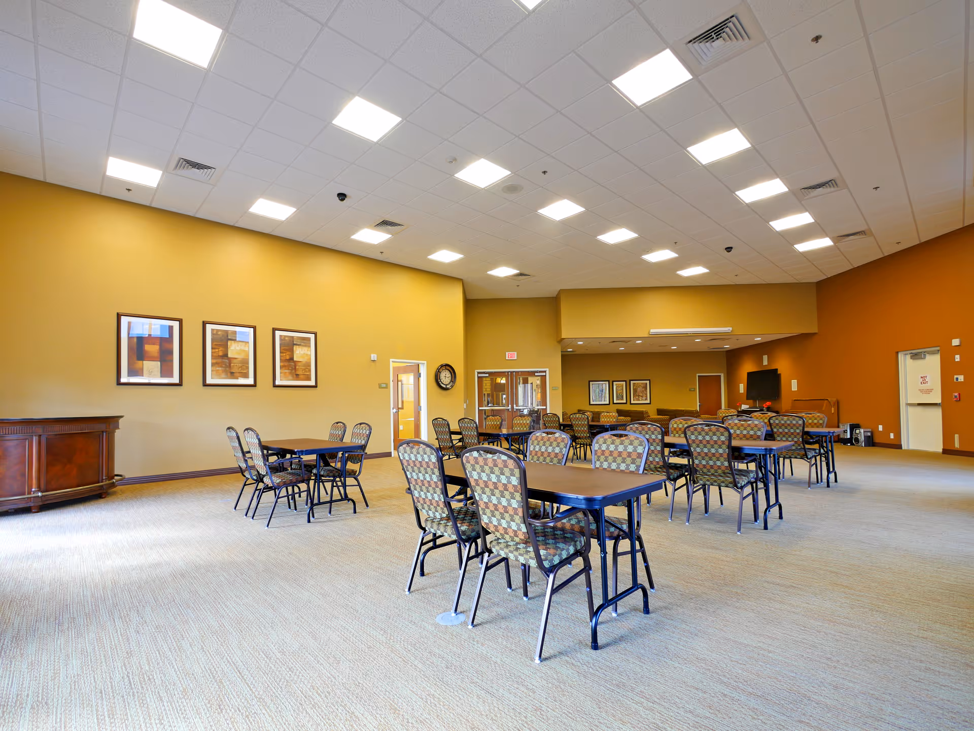 A spacious, well-lit common area with multiple tables and patterned chairs arranged neatly. The walls are painted in warm tones of yellow and orange, adorned with framed artwork. The ceiling has recessed lighting and air vents. There is a wooden cabinet on the left side and a clock on the wall near a door. The room appears clean and organized, suitable for group activities or dining.