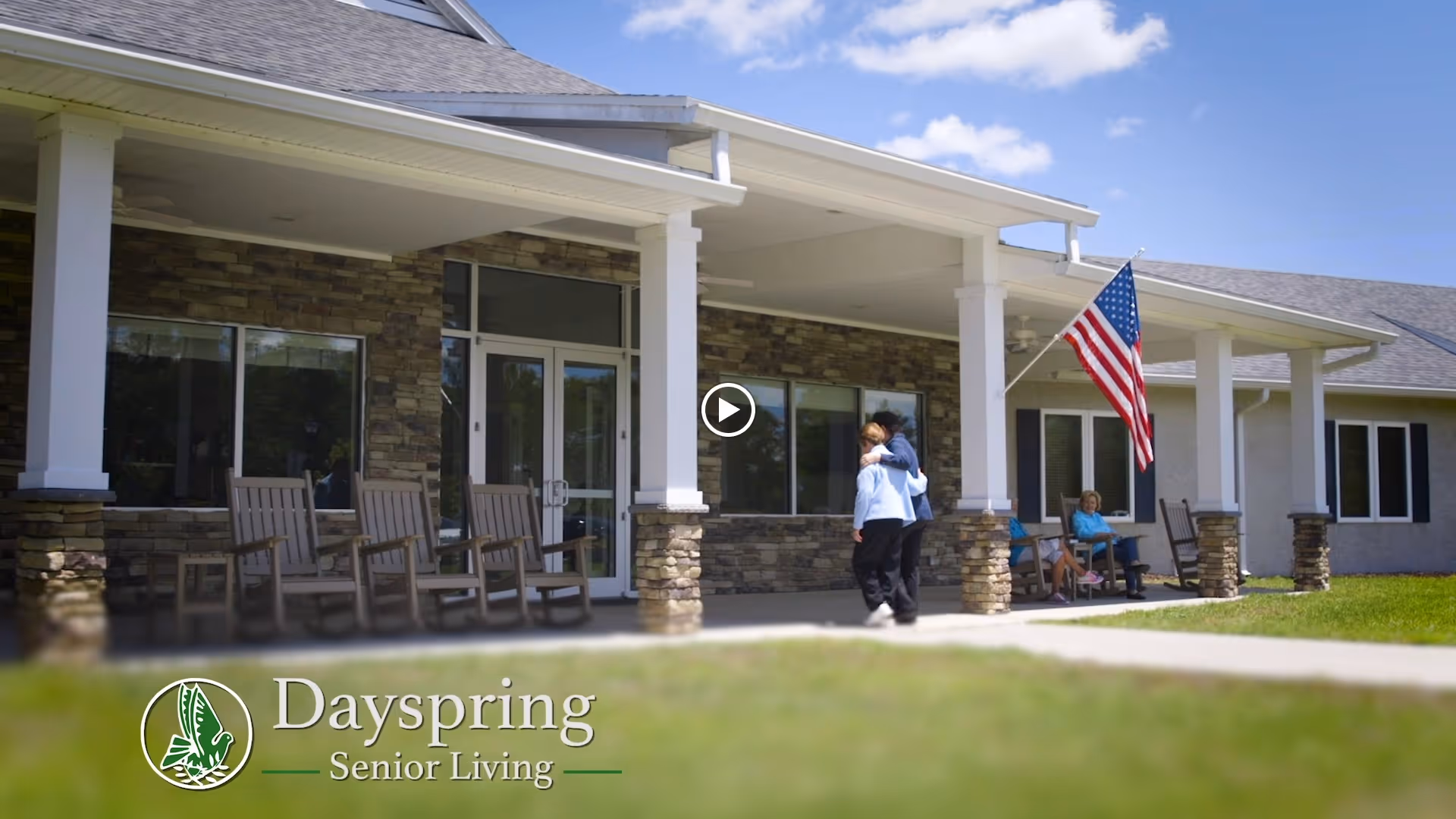Front entrance of a senior living facility with a covered porch, rocking chairs, an American flag, and people on the walkway.