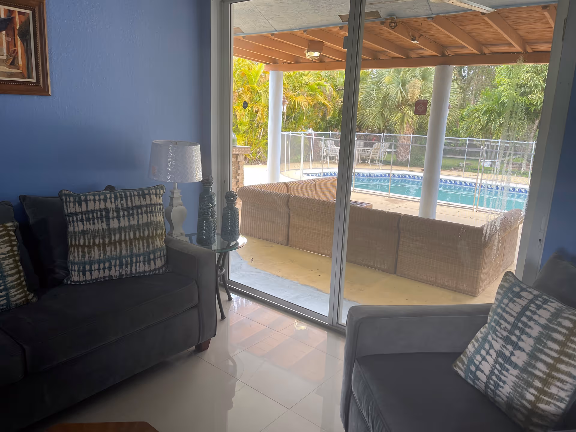 Living room with sofas and patterned pillows facing a sliding glass door that opens to a covered patio and a swimming pool.