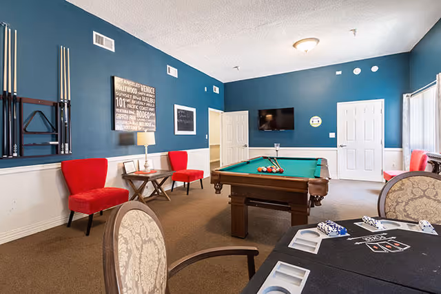 A recreational room with a pool table in the center, a wall-mounted TV, two red chairs with a small table and lamp between them, a rack holding pool cues, and a card table with chairs in the foreground. The walls are painted blue with white wainscoting and there are two white doors and a window with blinds.