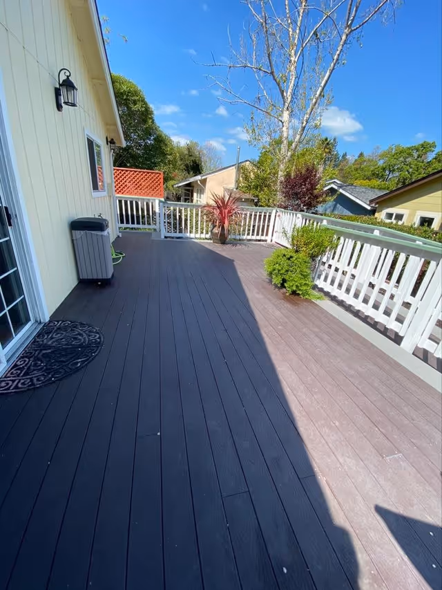 Outdoor deck area attached to a yellow building with white railing, potted plants, a trash bin, and a black doormat under a clear blue sky.