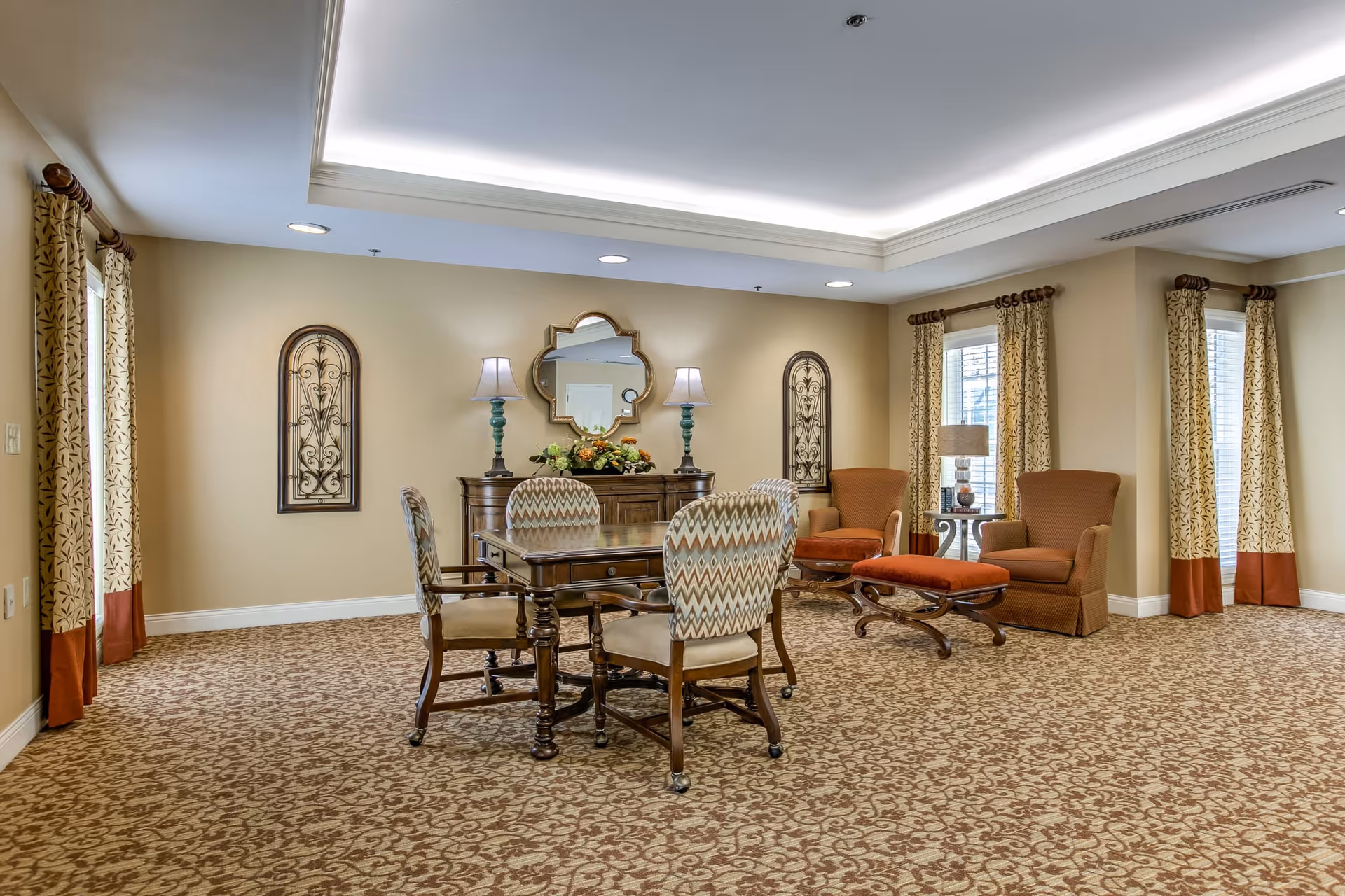 A well-lit room with beige walls and patterned carpet featuring a wooden table with four upholstered chairs in the center. Two armchairs with matching ottomans are positioned near windows with patterned curtains. A wooden sideboard with two lamps and a decorative mirror hangs on the wall behind the table.