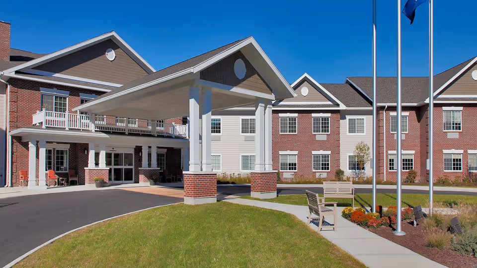 Exterior view of The Landing of Poughkeepsie senior living facility showing a large covered entrance with white columns and brick bases, a driveway, green lawn, benches, and flagpoles under a clear blue sky.
