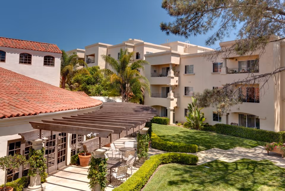 Outdoor courtyard area of La Vida Del Mar facility featuring a well-maintained lawn, trimmed hedges, palm trees, and a building with balconies. There is a wooden pergola with climbing plants and outdoor seating underneath. The sky is clear and blue.