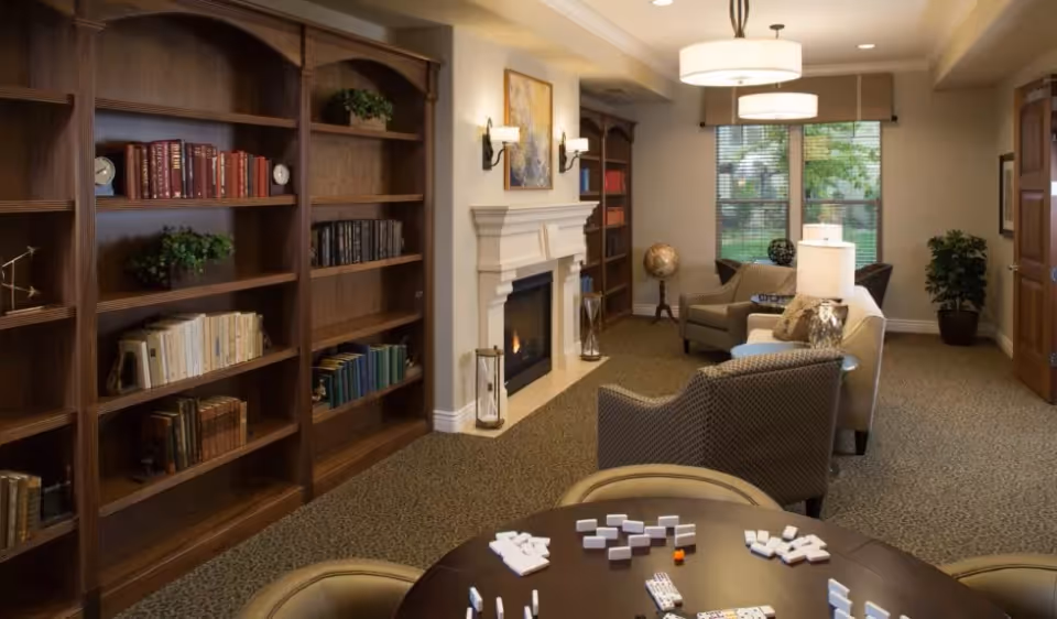 Cozy common living room with wooden bookshelves, a fireplace, seating area, window, and a game table with tiles.