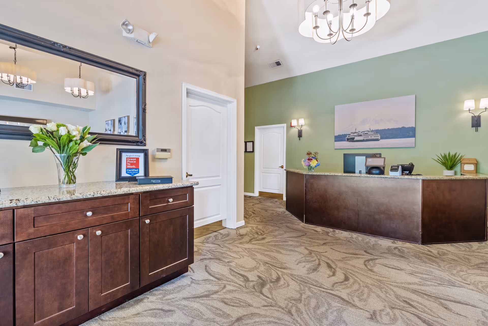 Reception area of Aegis Living Kent facility with a dark wood front desk, green accent wall, a large framed photo of a ferry and mountain, two white doors, a chandelier, wall sconces, and a cabinet with a vase of white flowers and a framed certificate.
