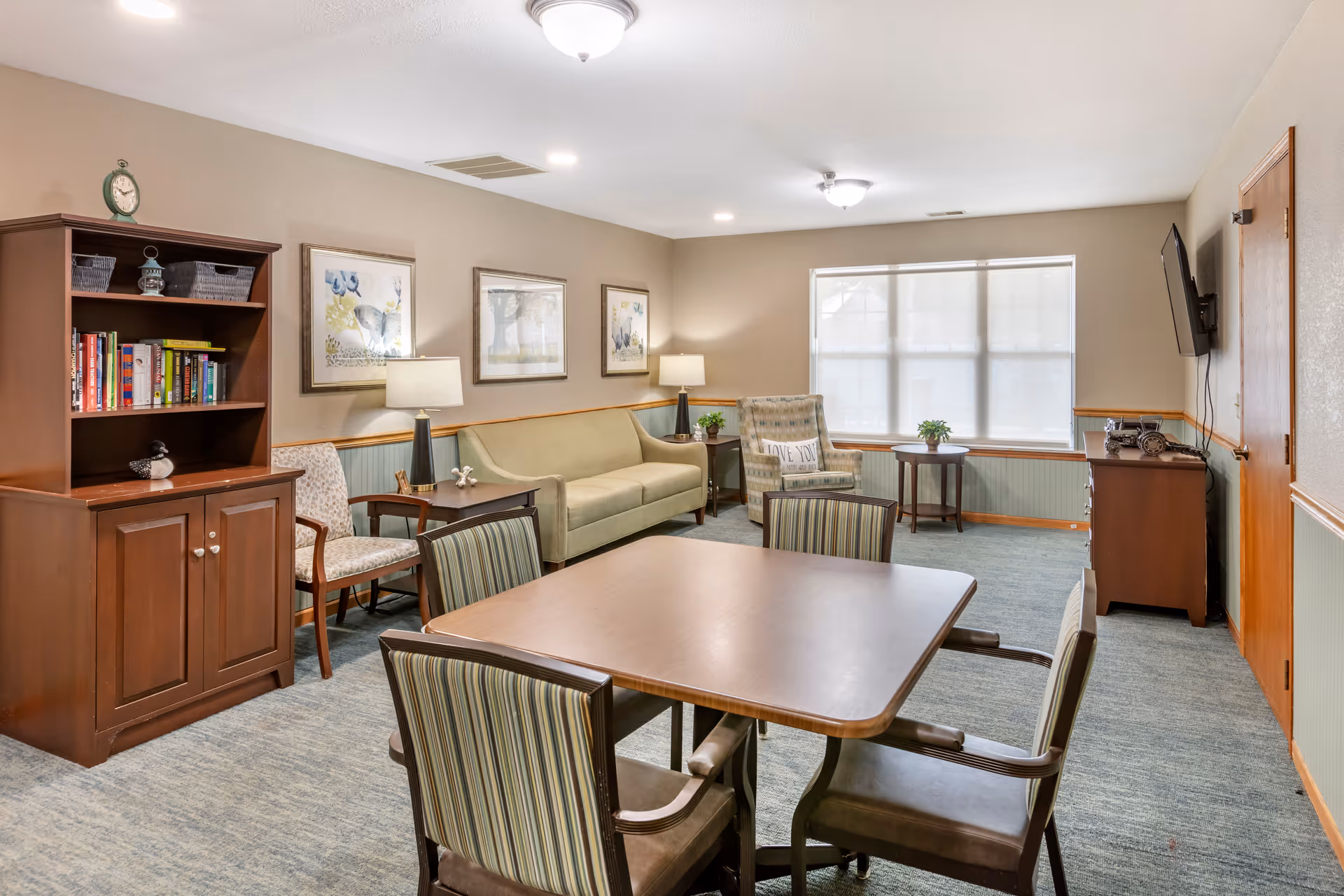 A cozy living room area with a square wooden table surrounded by four striped upholstered chairs in the foreground. In the background, there is a green sofa, an armchair with a 'Love You More and More' pillow, two side tables with lamps, and a wooden bookshelf with books and decorative items. The room has beige walls with three framed pictures, a large window with blinds, and a wall-mounted TV on the right side.