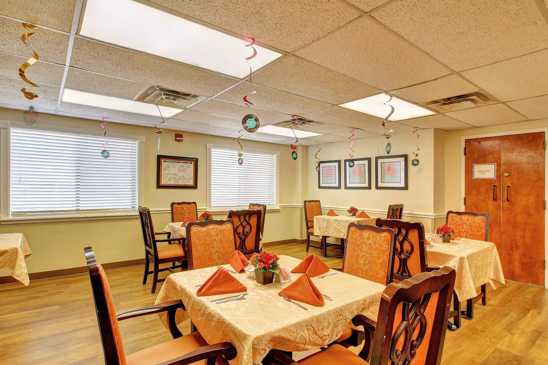 Dining room with tables set with folded napkins and floral centerpieces, upholstered wooden chairs, hanging ceiling streamers, and framed artwork on the walls.