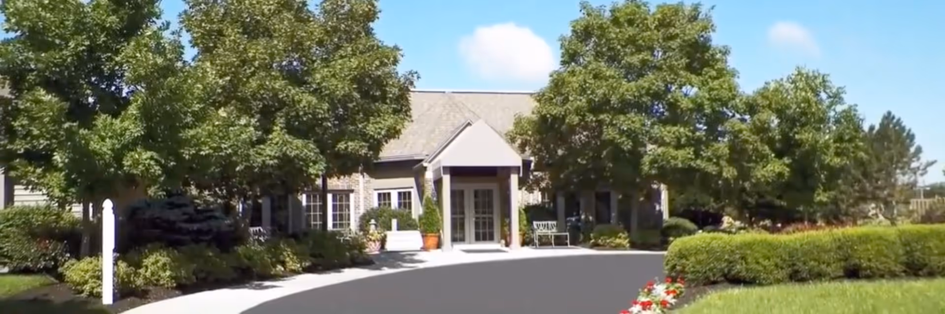 Single-story building entrance with a covered doorway, flanked by trees, shrubs, and a paved driveway.
