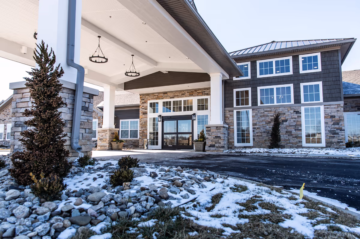 Exterior view of a senior living facility entrance with stone and wood siding, large windows, and a covered driveway. Snow is visible on the ground and landscaping rocks.