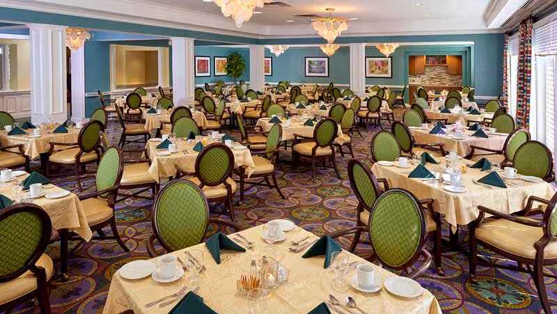 A spacious dining room with multiple round tables covered with beige tablecloths, each set with white plates, cups, silverware, and green folded napkins. The chairs have green cushioned backs and seats with dark wooden frames. The room features patterned carpet, blue walls with white trim, framed artwork, and large windows with blinds allowing natural light to enter.