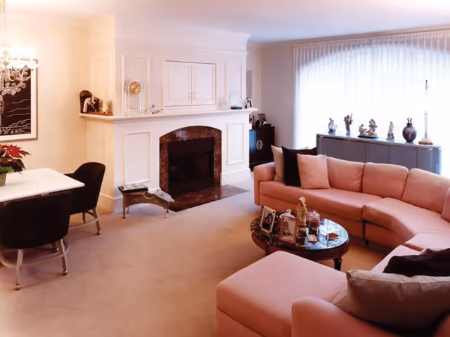 A cozy living room with a pink sectional sofa and matching ottoman, a round glass coffee table with framed photos and decorative items, a white fireplace with a dark marble hearth, a dining table with black chairs, and a large window with sheer white curtains letting in natural light.