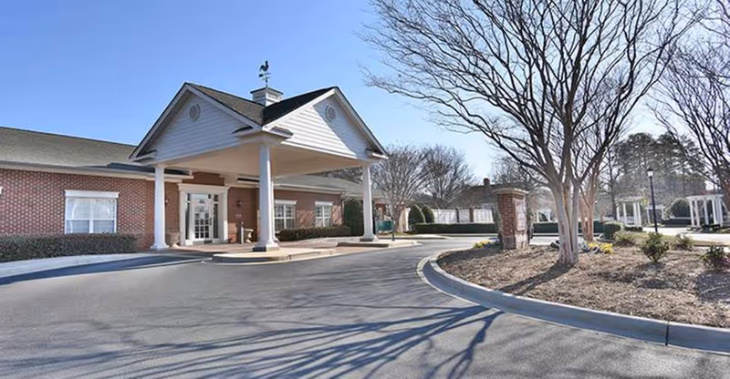 Exterior view of a senior living facility entrance with a covered drop-off area supported by white columns, red brick walls, and a paved driveway. Leafless trees and landscaped areas are visible around the driveway under a clear blue sky.