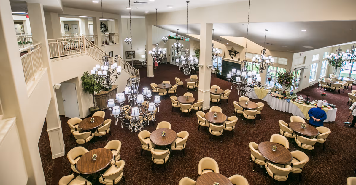 Spacious dining room with multiple round wooden tables surrounded by beige cushioned chairs. Elegant chandeliers hang from the ceiling, illuminating the room. There is a buffet table with food and a person standing nearby. The room has large windows allowing natural light to enter and a staircase leading to an upper level.