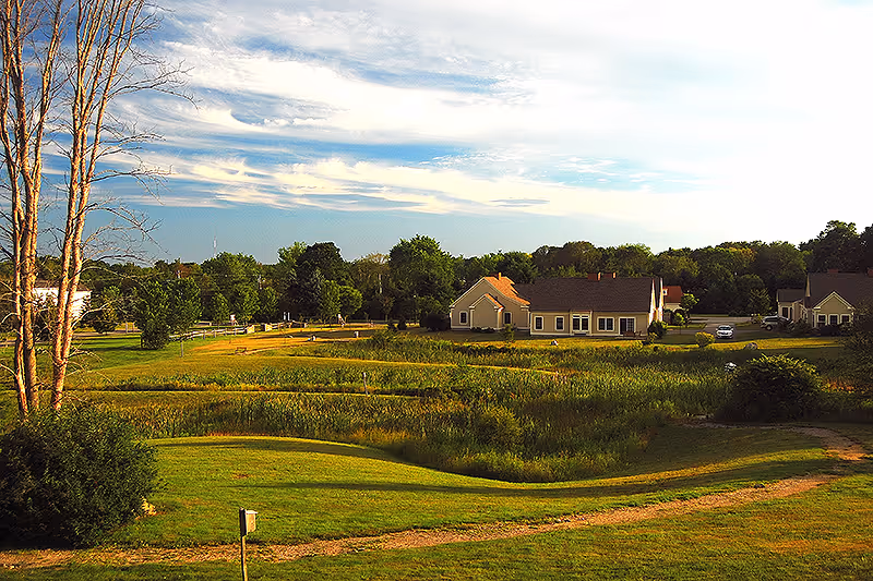 A scenic outdoor view of a senior living facility with green lawns, trees, and several single-story buildings under a partly cloudy sky.