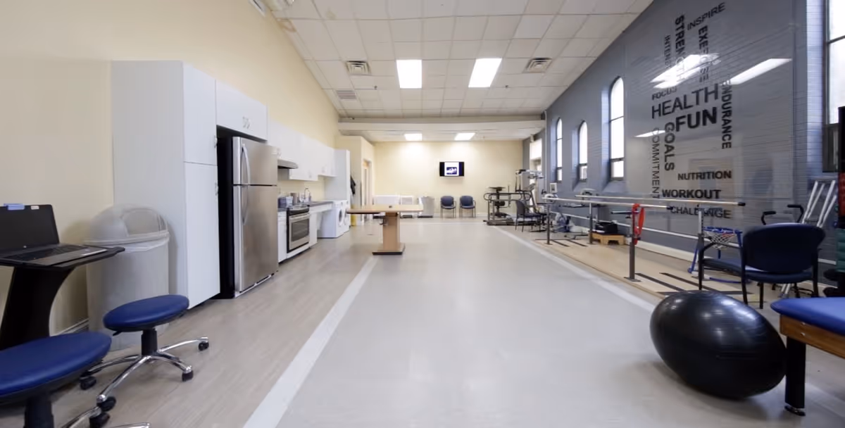 A spacious rehabilitation room with a kitchenette on the left side including a refrigerator, stove, and cabinets. On the right side, there are parallel bars for walking exercises, exercise equipment, a large exercise ball, and chairs. The far wall has a mounted TV and three chairs beneath it. The room has a high ceiling with fluorescent lighting and several arched windows on the right wall. Motivational words like HEALTH, FUN, WORKOUT, and NUTRITION are displayed on the right wall.