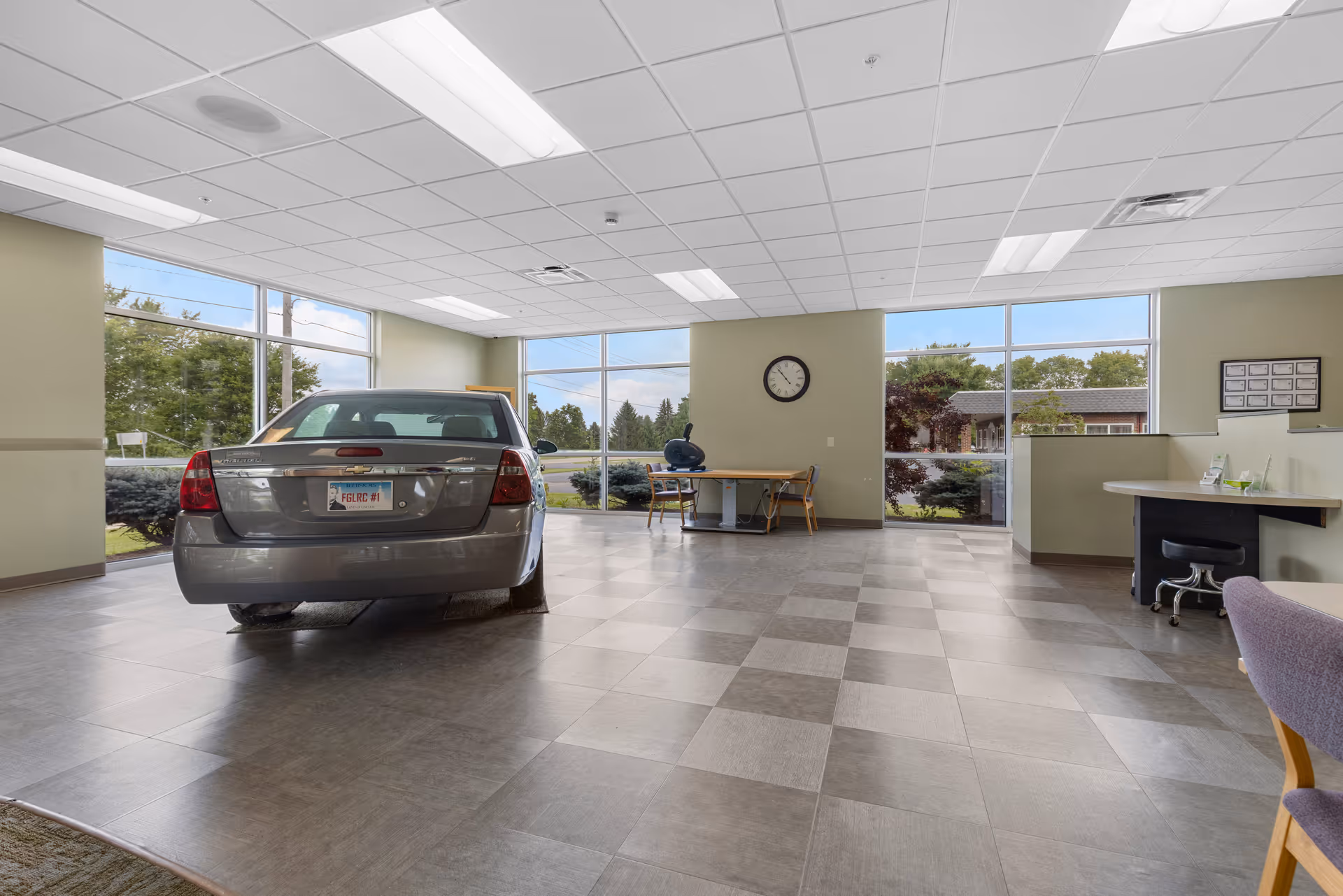 A spacious room with large windows showing an outdoor view of trees and a building. Inside the room, there is a silver car parked on a tiled floor. The room has a drop ceiling with fluorescent lights, a wall clock, a desk with a chair, and another workstation with a stool. The walls are painted light green.