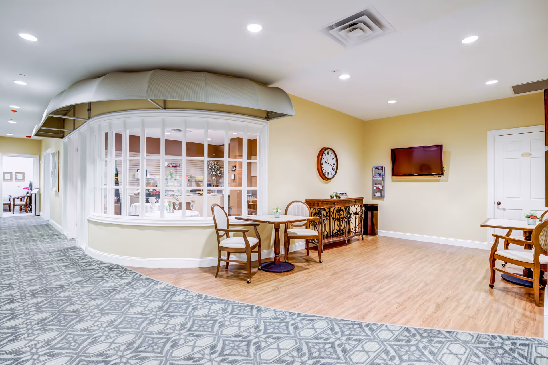 Interior view of a senior living facility hallway with a seating area featuring wooden chairs and tables. There is a large windowed room on the left side with white framing and a beige awning above it. The floor has a patterned carpet in the hallway and wood flooring in the seating area. A wall clock and a mounted TV are visible on the beige walls.