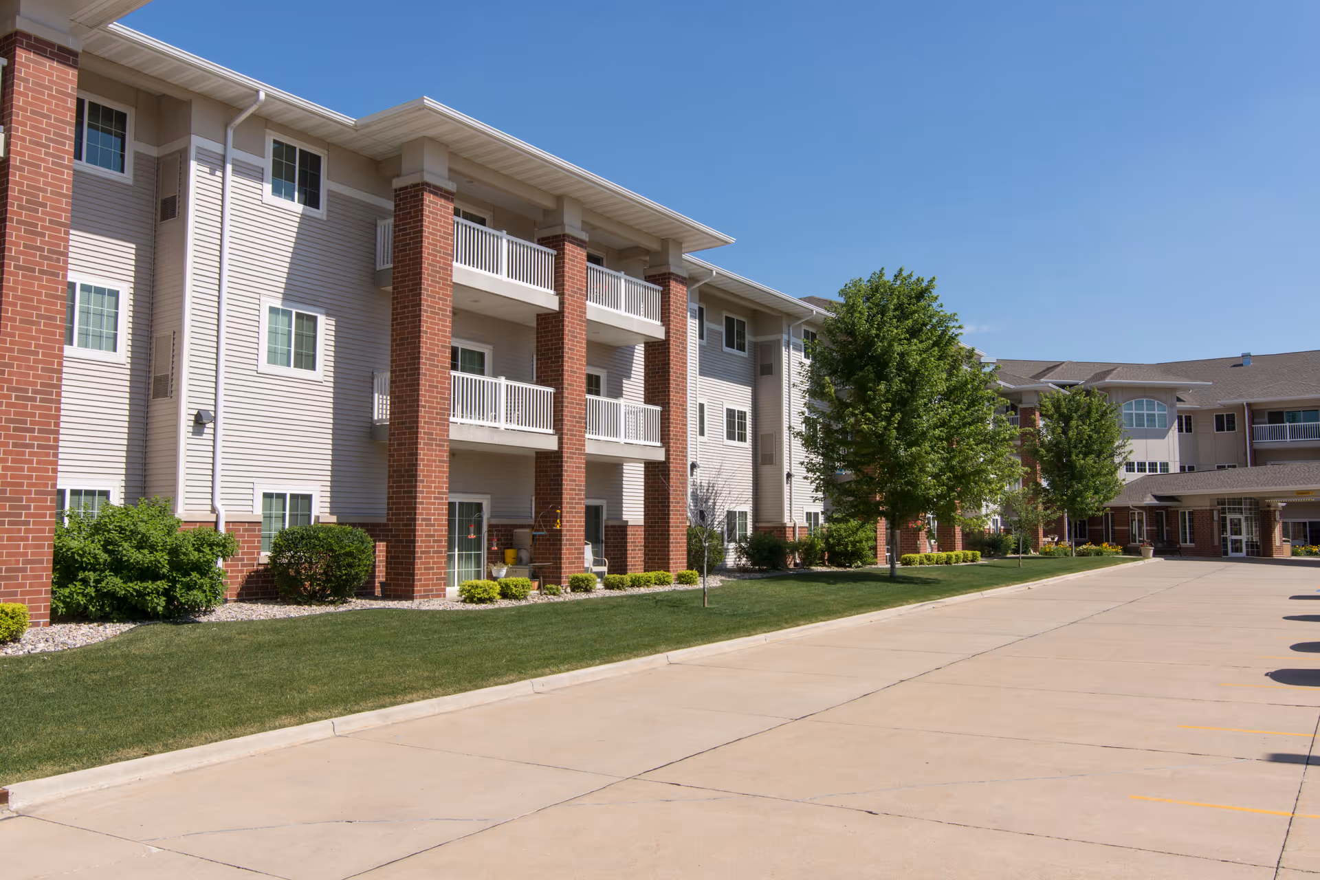 Multi-story retirement community building exterior with balconies, landscaped lawn, trees, and a wide driveway.