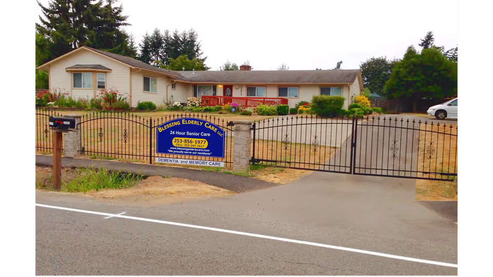 A single-story residential building behind a black iron gate with a sign that reads 'Blessing Elderly Care LLC, 24 Hour Senior Care, Dementia and Memory Care'. The building has beige siding, several windows, a red front porch railing, and is surrounded by some greenery and trees. A mailbox is visible near the gate, and a white car is parked on the right side of the property.
