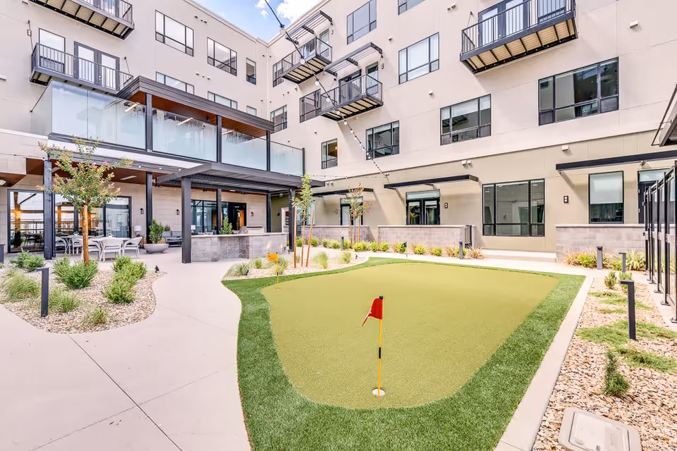 Outdoor courtyard area of a senior living facility with a small putting green featuring a red flag, surrounded by landscaped plants and trees. The courtyard is enclosed by a multi-story building with balconies and large windows. There is a covered seating area with tables and chairs on the left side.