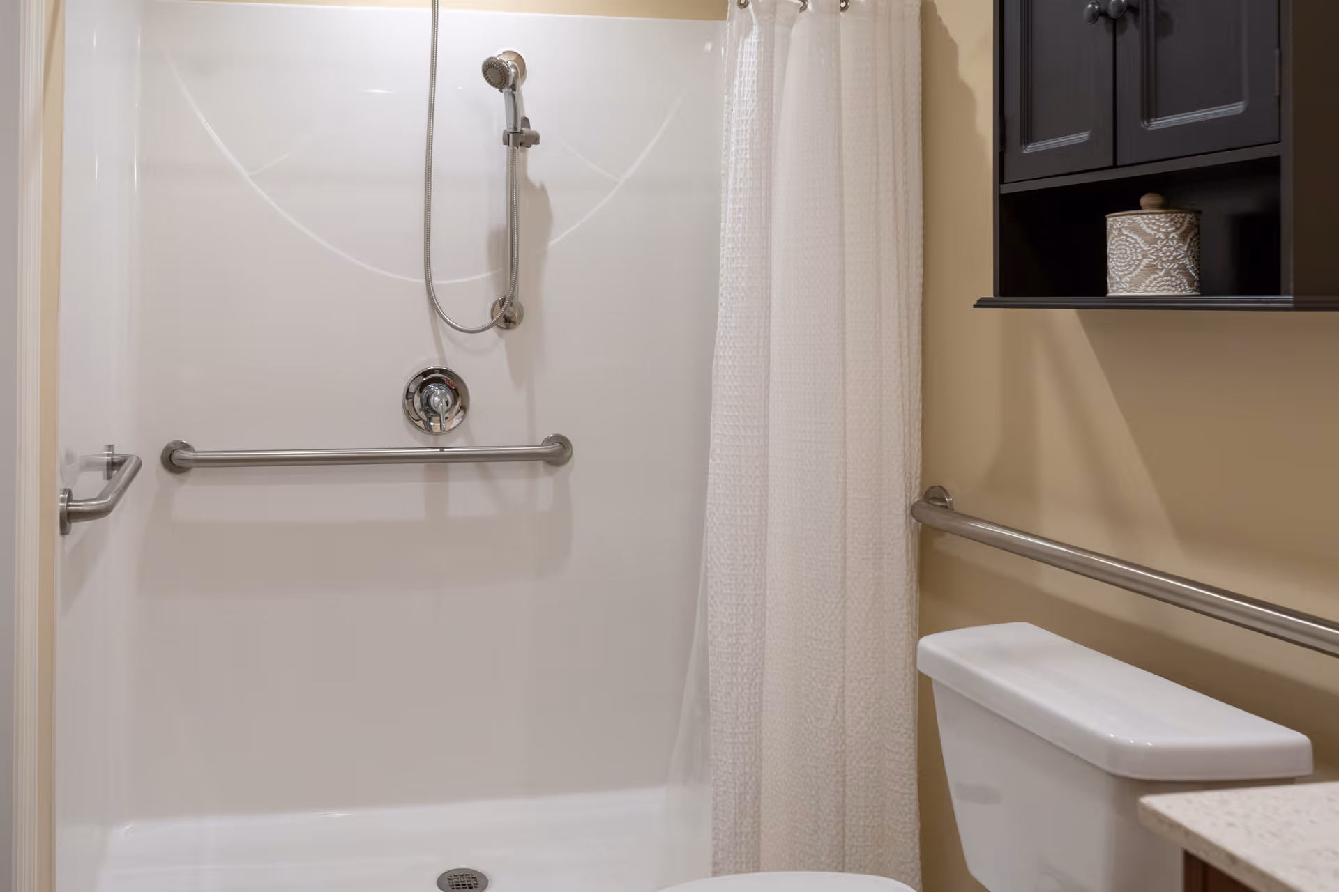 A bathroom with a white shower area featuring a handheld showerhead and a horizontal grab bar. There is a white shower curtain, a toilet with a horizontal grab bar beside it, and a dark wall-mounted cabinet with a decorative container on the shelf.