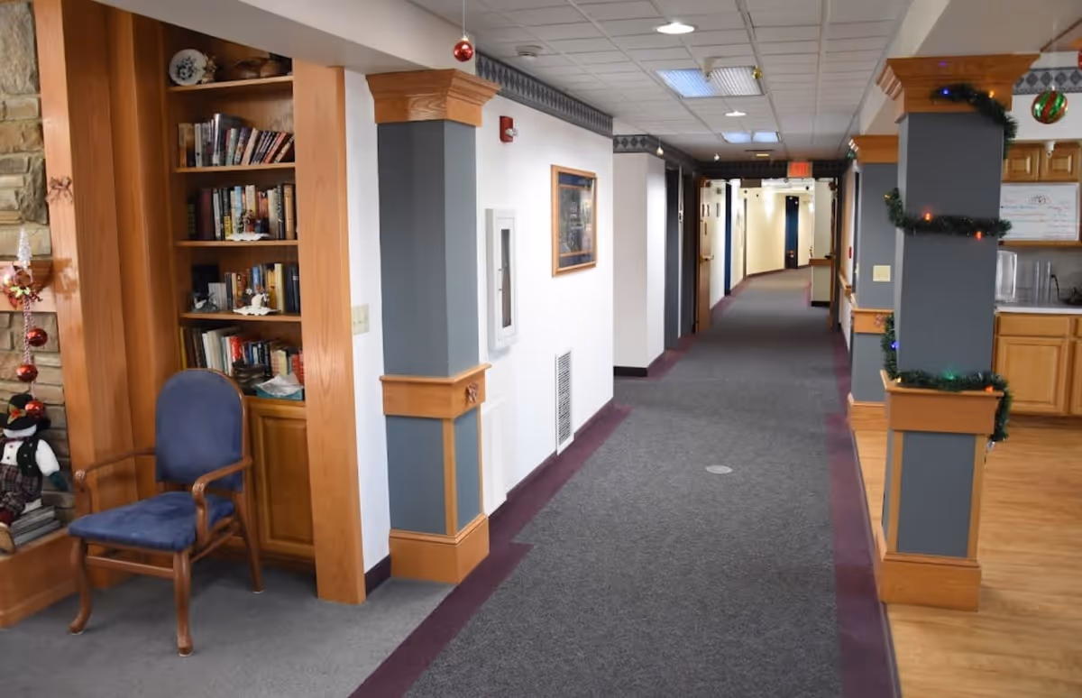 Interior hallway of Cedar Vale Assisted Living with carpeted floor and wooden trim. On the left side, there is a built-in bookshelf filled with books and a blue cushioned wooden chair. The hallway is decorated with holiday ornaments and garlands, and there is a kitchen area visible on the right side.