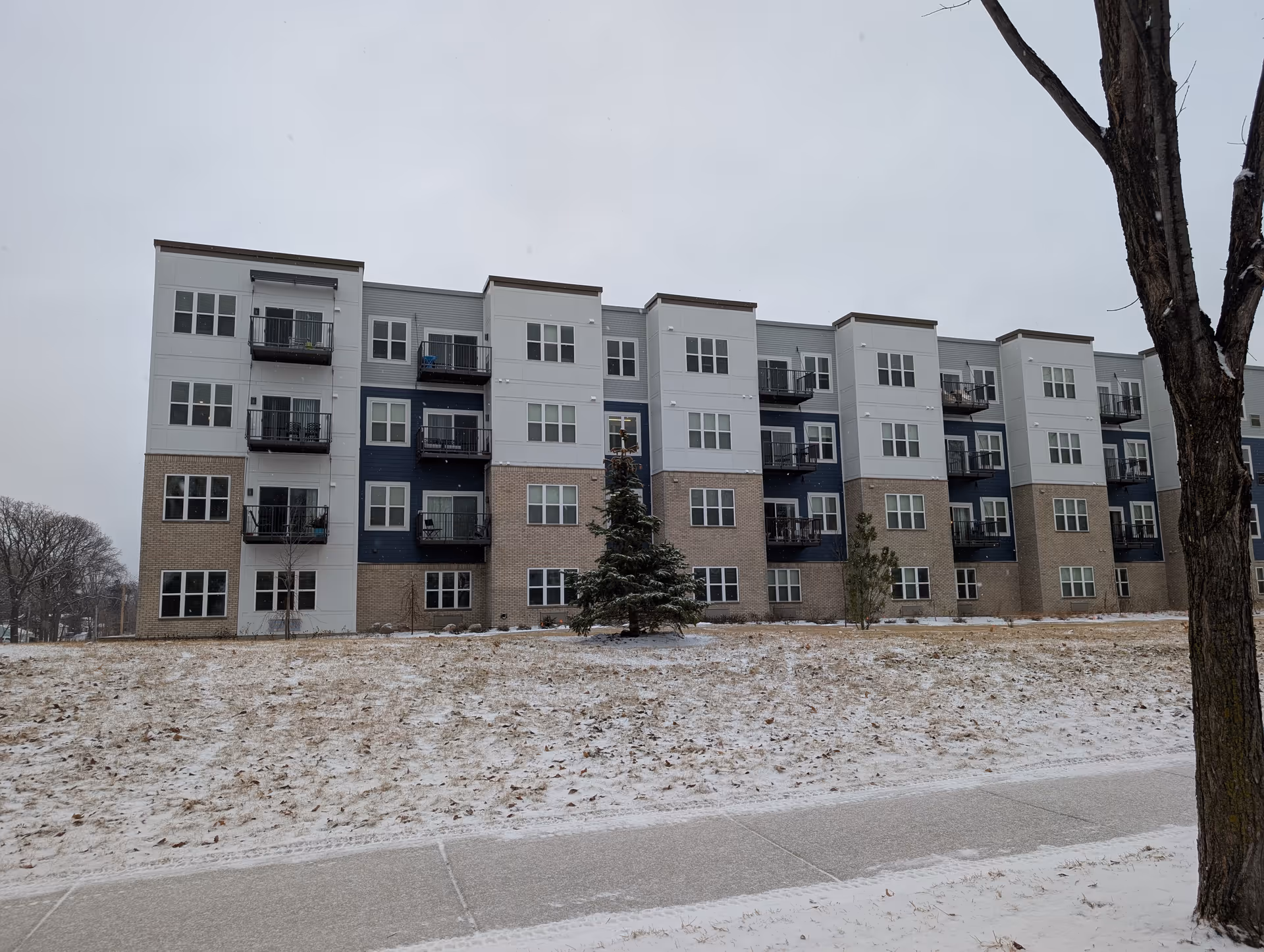 Multi-story modern apartment building with balconies viewed across a snow-dusted lawn and sidewalk.
