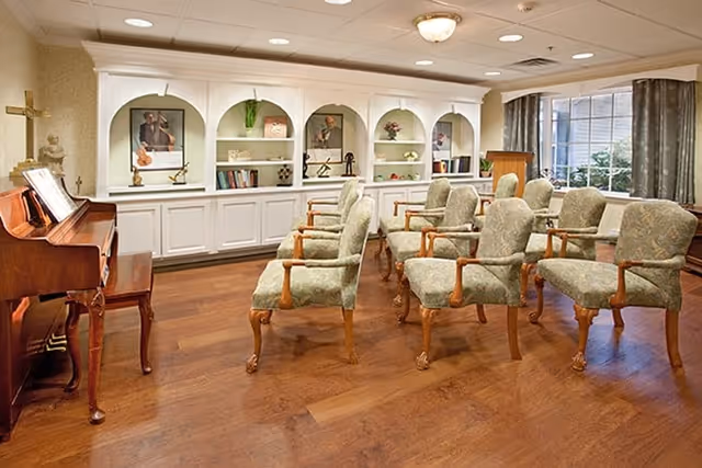 A small meeting or music room with wooden flooring, upholstered chairs arranged in rows, a wooden piano on the left, and built-in white shelves with decorative items and framed pictures on the back wall. A window with curtains is on the right side.