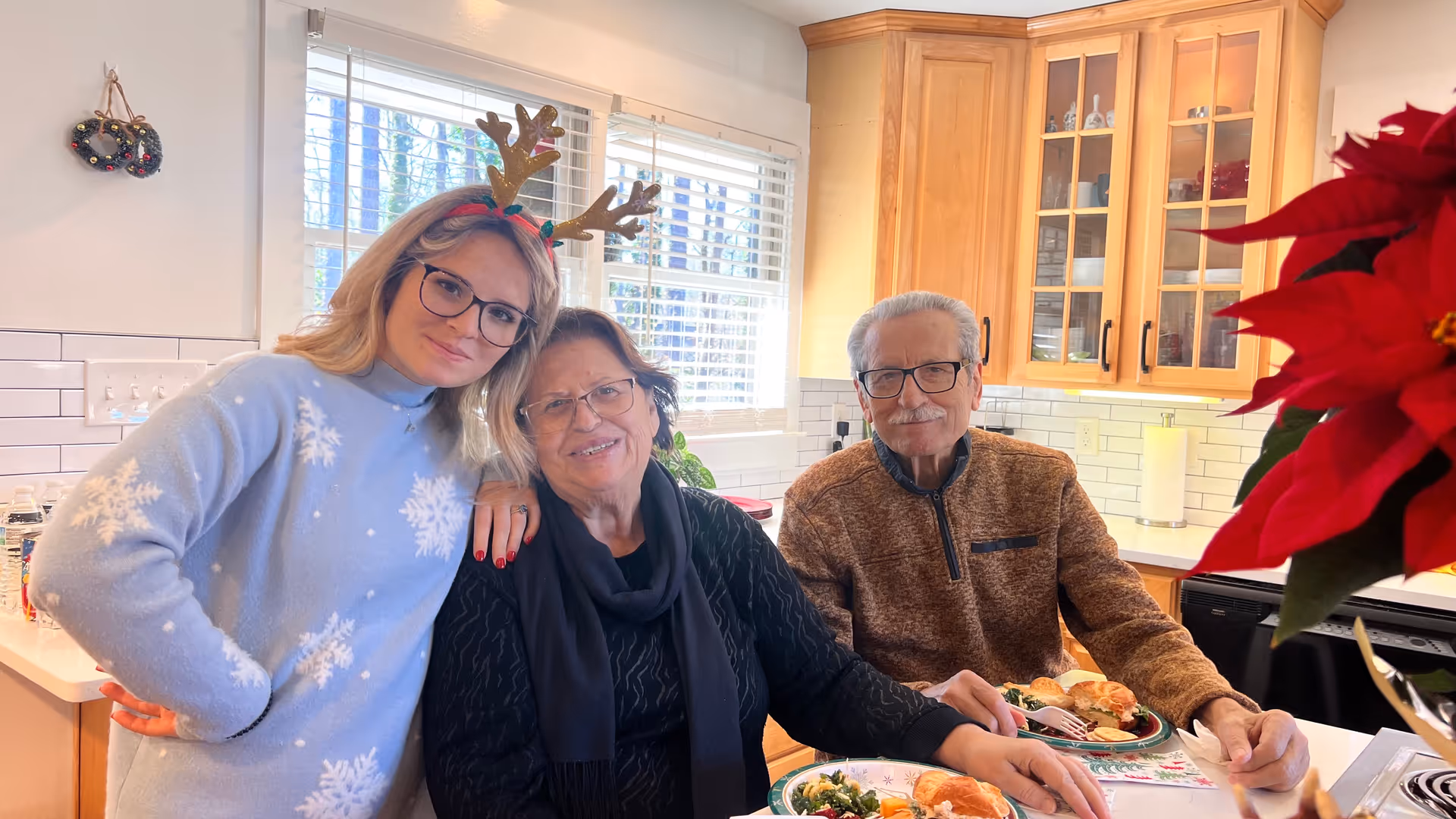 A young woman wearing reindeer antlers and a light blue sweater with snowflake patterns stands next to an elderly woman and an elderly man seated at a kitchen counter with plates of food. The kitchen has wooden cabinets, white subway tile backsplash, and a window with blinds. A red poinsettia plant is visible in the foreground.