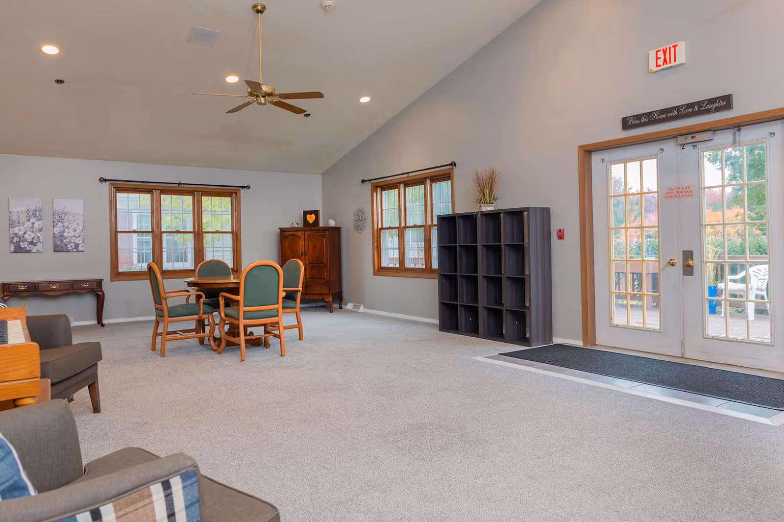 A spacious senior living common area with beige carpet, light gray walls, and wooden trim around windows and doors. The room features a round wooden table with four green cushioned chairs, a wooden cabinet, a black cubby shelf, and a piano with a gray armchair nearby. French doors lead outside, and a ceiling fan with lights hangs from the ceiling. A sign above the door reads 'Bless this Home with Love & Laughter.'