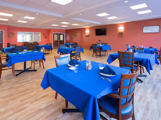 A spacious dining room with multiple tables covered in blue tablecloths, each set with napkins, utensils, and small centerpieces. The room has wooden chairs, wood-patterned flooring, coral-colored walls, ceiling lights, a wall-mounted TV, and decorative wall art.
