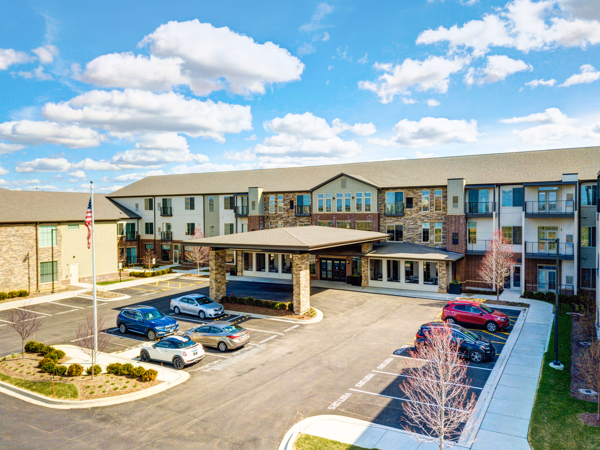 Exterior view of The Oaks at Algonquin Senior Living facility showing a three-story building with a covered entrance, several parked cars, a flagpole with an American flag, and a partly cloudy blue sky.