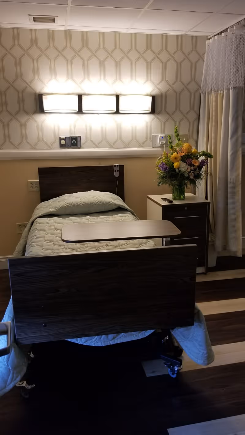 A nursing facility patient room with a single adjustable bed, overbed table, nightstand topped with a vase of flowers, and wall lighting.