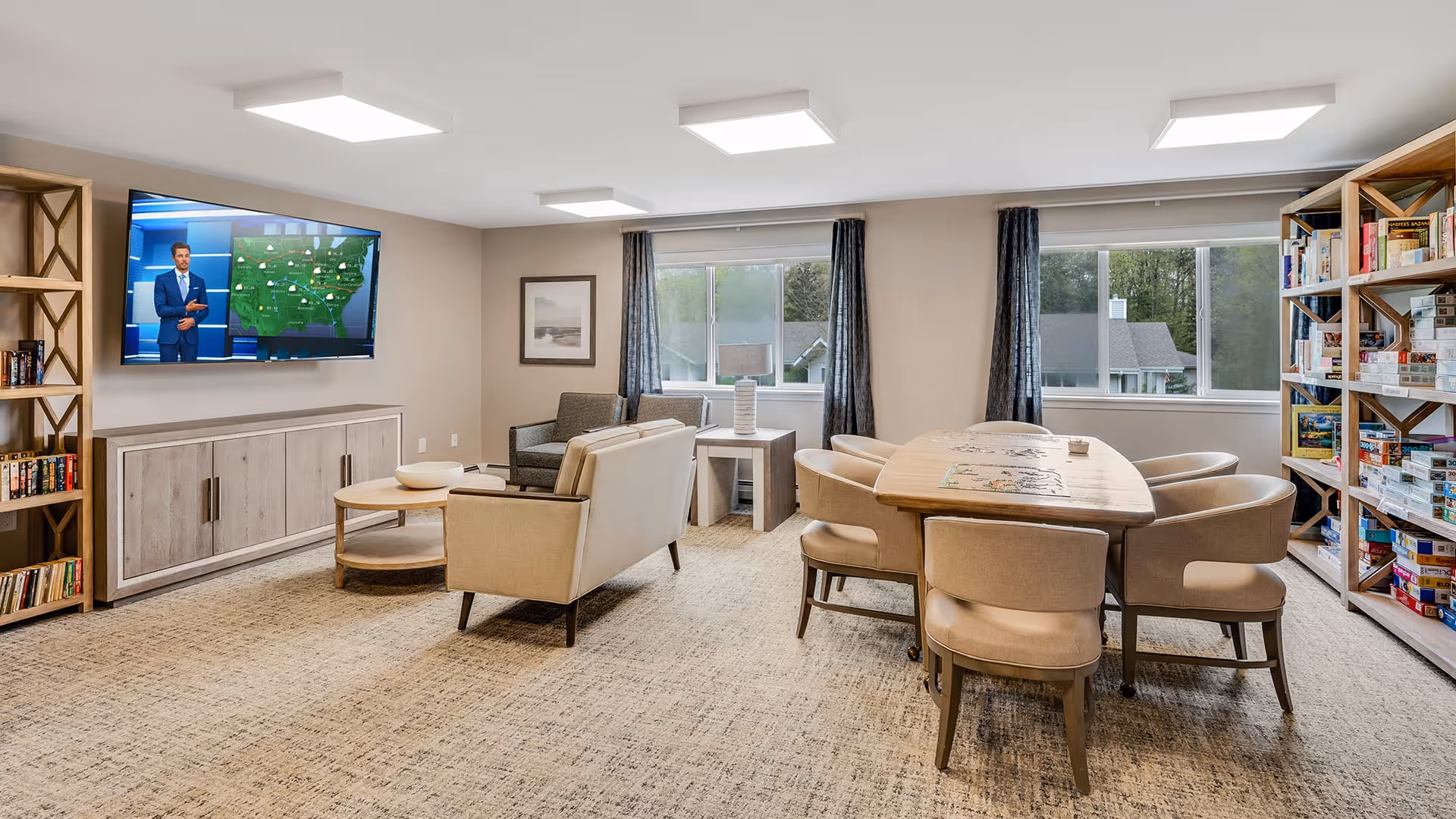 A bright and spacious common area with beige carpeting, featuring a seating area with two armchairs and a loveseat facing a wall-mounted TV displaying a weather forecast. There is a wooden coffee table in front of the seating, and a large wooden table with six upholstered chairs arranged around it. Bookshelves filled with books and board games line the walls. Large windows with dark curtains let in natural light and offer a view of trees and houses outside.